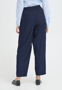 Tumesinised laia lõikega püksid, millel on elastne vöökoht, tagataskud ja sile tekstuur, kombineeritud mustade lamedate kingadega.