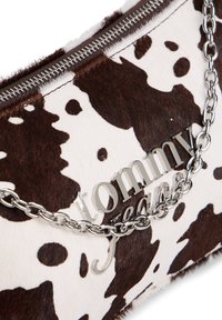 Plic cu imprimeu alb și maro de vacă, detalii cu lanț argintiu și un logo lustruit. Are închidere cu fermoar în partea de sus.