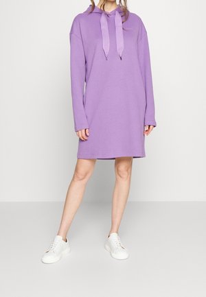 Person, die ein langärmeliges fliederfarbenes Hoodie-Kleid mit breiten Kordeln und weißen Turnschuhen trägt, steht vor einem einfachen hellen Hintergrund.