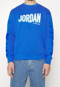 Blå sweatshirt med avslappnad passform, ribbade ärmslut och texturerade detaljer på ärmarna; har "JORDAN" i fet vit text på framsidan.