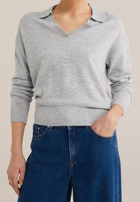 Vaalea harmaa neulepusero, jossa on V-pääntie ja käännekaulus, tiukat hihansuut ja ribattua helmaa, yhdistettynä korkeavyötäröisiin sinisiin farkkuihin.