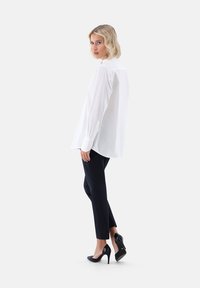 Weißes langärmliges Hemd mit Knopfleiste in lockerer Passform, kombiniert mit navy-farbenen, schmal geschnittenen Hosen und schwarzen High Heels. Glatte Textur, klassischer Kragen.