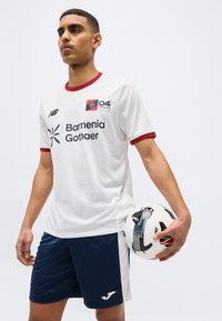 Νεαρός ποδοσφαιριστής με λευκή και κόκκινη φανέλα και σκούρο μπλε σορτς κρατώντας μια λευκή και μαύρη μπάλα ποδοσφαίρου, σε μονόχρωμο φόντο.