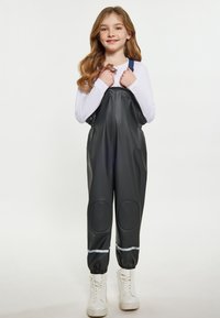 Juodi nepralaidūs overalai su priekiniais užtrauktukais, reguliuojamomis mėlynomis petnešomis, kelio įdėklais ir atšvaitinėmis detalėmis, derinami su baltais aukštakulniais sportbačiais.