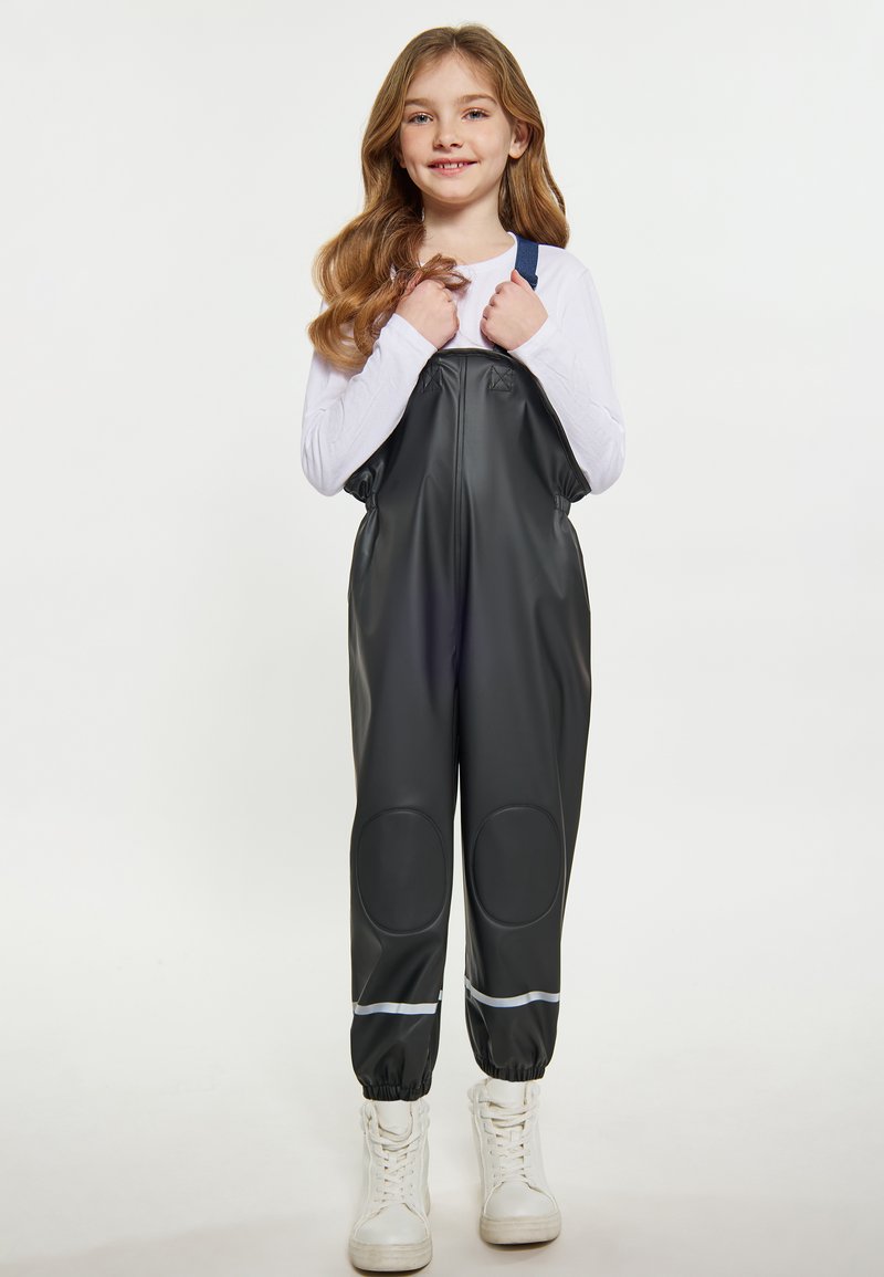 Juodi nepralaidūs overalai su priekiniais užtrauktukais, reguliuojamomis mėlynomis petnešomis, kelio įdėklais ir atšvaitinėmis detalėmis, derinami su baltais aukštakulniais sportbačiais.
