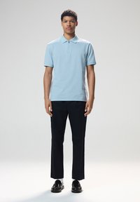 Polo azzurro chiaro con colletto con zip, tessuto texture, abbinato a pantaloni neri e scarpe nere lucide, in piedi su uno sfondo neutro.