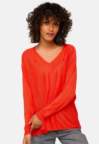 Roter, leichter V-Ausschnitt-Pullover mit langen Ärmeln, lässiger Passform und strukturiertem Finish. Kombiniert mit lässigen grauen Jeans.