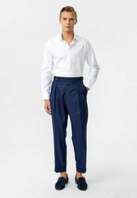 Weißes Langarmhemd kombiniert mit dunkelblauer Hose mit vorderen Falten und seitlichen Knöpfen. Die Hose endet knöchellang.