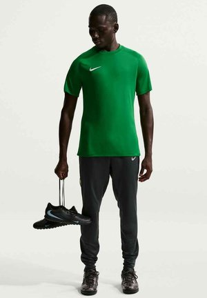 Uomo con maglietta verde Nike e pantaloni neri che tiene in mano scarpe da calcio nere Nike per i lacci su sfondo bianco.