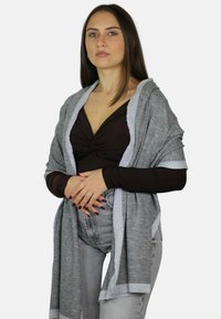 Cardigan grigio con una texture morbida, caratterizzato da un bordo argentato lungo i bordi. Indossato sopra una maglia scura, abbinato a jeans chiari.