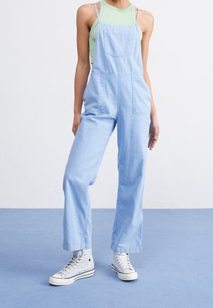 Vaaleansiniset farkkuhaalari, jossa on etutasku, leveät lahkeet ja teksturoitu kangas. Yhdistetty mintunvihreään hihattomaan toppiin ja valkoisiin korkeavartisiin tennareihin.