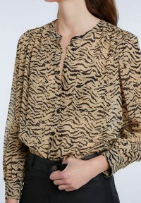 Beige Bluse mit schwarzem Tigerstreifenmuster, langen Ärmeln, Knopfleiste und einer Brusttasche, hergestellt aus leichtem, transparentem Stoff.