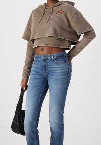 Hnědá zkrácená mikina s červeným logem, spárovaná se světlými modrými džíny. Modelka drží černou pletenou kabelku.