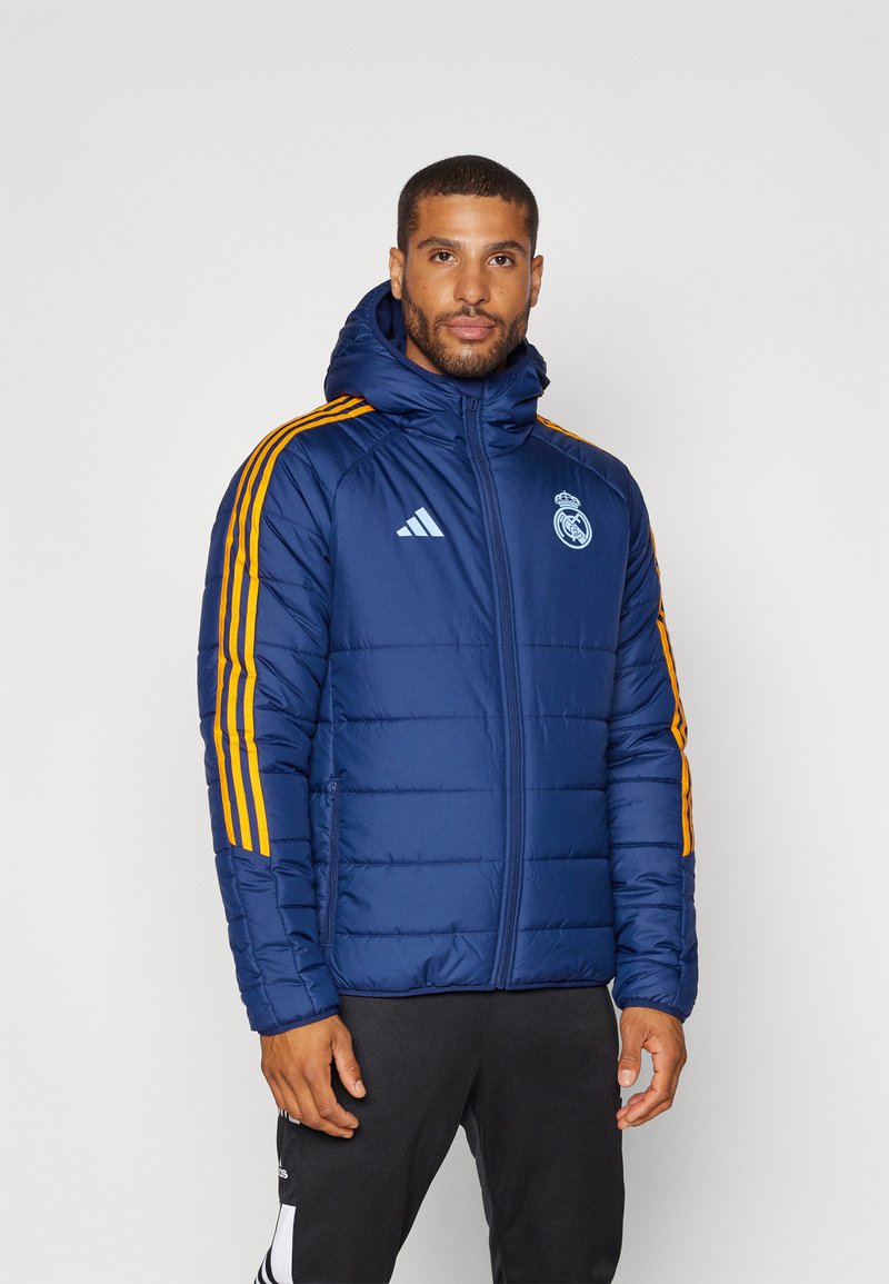 Giacca a vento blu navy con cappuccio, caratterizzata da strisce gialle sulle spalle, un logo ricamato sul petto e una texture trapuntata. Chiusura frontale con zip.