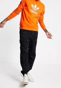 Oranžová mikina s bílým logem Adidas, doplněná černými cargo kalhotami s kapsami a bílými sportovními botami.