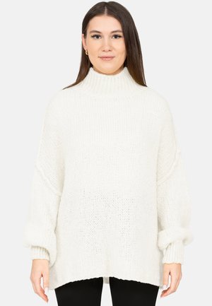 Femme aux longs cheveux bruns portant un pull à col roulé blanc en maille épaisse et un pantalon noir sur un fond uni.