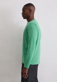 Koszulka z długim rękawem w jednolitym jasnym zielonym kolorze, wykonana z bawełny, z okrągłym dekoltem. Noszona z ciemnymi dżinsami, prezentowana na neutralnym tle.