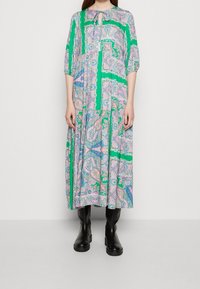 Dlouhé zelené květinové šaty s paisley vzorem, s výstřihem ve tvaru klíče a tříčtvrtečními rukávy. Doplněné černými vysokými botami po kolena.