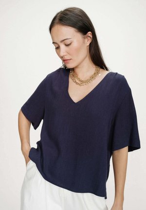 Femme aux longs cheveux foncés portant un haut bleu marine à col en V et manches courtes, un pantalon blanc et des colliers en chaîne d’or superposés, regardant vers le bas avec une main dans la poche.