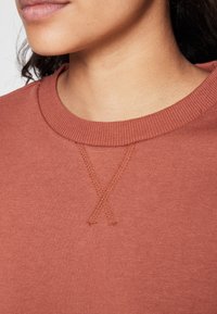 Primer plano del cuello y la parte inferior de la cara de una persona que lleva un sudadera de color óxido con cuello redondo acanalado y un detalle decorativo de costura en forma de "X".