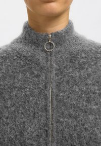 Gros plan d'une personne portant un pull zippé gris duveteux avec un tire-zip rond en argent au niveau du cou.
