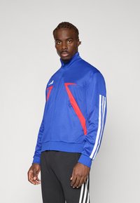 Blå sportsjakke med røde detaljer, høy krage og hvite striper på ermene, laget av et glatt og elastisk stoff.