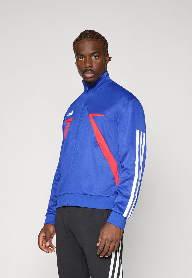 Blå sportsjakke med røde detaljer, høy krage og hvite striper på ermene, laget av et glatt og elastisk stoff.