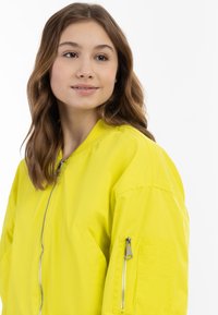 Giacca bomber gialla brillante con zip frontale, tasche laterali e tasca con zip sulla manica. Tessuto morbido e leggero, vestibilità comoda.