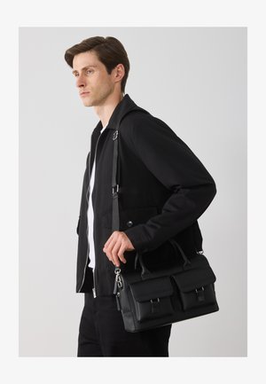 Svart lærveske med to frontlommer, metallklips, dobbel håndtak og justerbar skulderreim. Modellen har på seg en svart jakke og hvit skjorte.