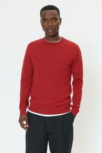 Pulover roșu tricotat, cu guler rotund și mâneci lungi. Purtați peste o cămașă albă, asortat cu pantaloni negri, având o textură subtilă.