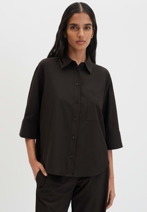 Vrouw met lang zwart haar die een losse zwarte blouse draagt met driekwart mouwen en bijpassende zwarte broek, hand in de zak.