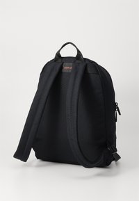 Mochila de tela negra con correas ajustables y acolchadas, un asa en la parte superior y cierre de cremallera. Presenta una pequeña etiqueta de logo roja en la parte delantera.