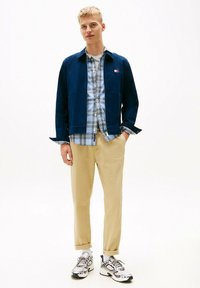 Chaqueta azul marino sobre una camisa de cuadros, pantalones beiges y zapatillas deportivas blancas. Las características incluyen una camisa con botones al frente y los puños de los pantalones enrollados.