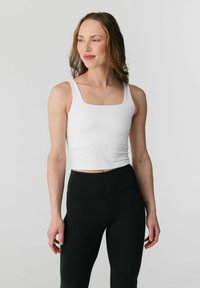 Vrouw met schouderlang bruin haar, gekleed in een witte mouwloze crop top met vierkante hals en hoge taille zwarte leggings, kijkt opzij.
