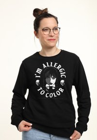 Donna con occhiali che indossa un felpa nera con una ritrattista gotica, teschi e la scritta "Sono allergica al colore", sorridendo mentre tiene il bordo.