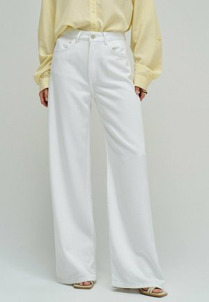 Femme portant un jean blanc taille haute à jambes larges, un chemisier jaune pâle, et des sandales beiges à bouts ouverts, debout devant un fond neutre.