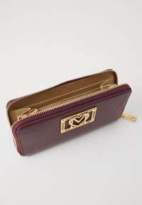 Dark burgundy leather wallet with gold-tone hardware, featuring a gold logo accent, zip closure, and beige interior.