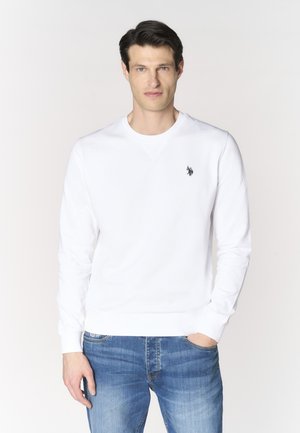 Uomo che indossa una felpa bianca semplice a maniche lunghe con piccolo logo ricamato sul petto, abbinata a jeans blu, in piedi su sfondo bianco.