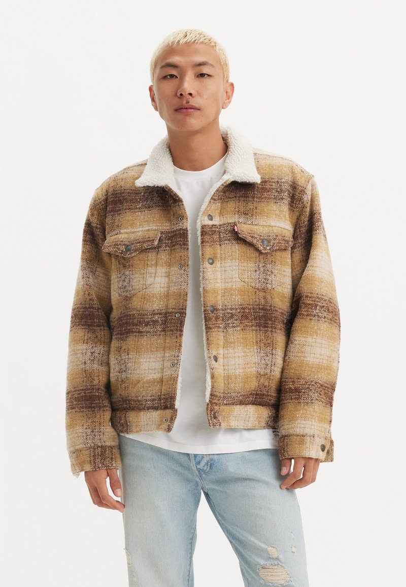 Chaqueta de cuadros marrón y amarilla con cuello sherpa, que presenta bolsillos en el pecho y cierres de botones. Llevada sobre una camiseta blanca.