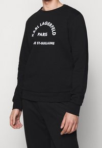 Persona con felpa nera a maniche lunghe con la scritta bianca "KARL LAGERFELD PARIS RUE ST-GUILLAUME" sul davanti, abbinata a pantaloni neri.