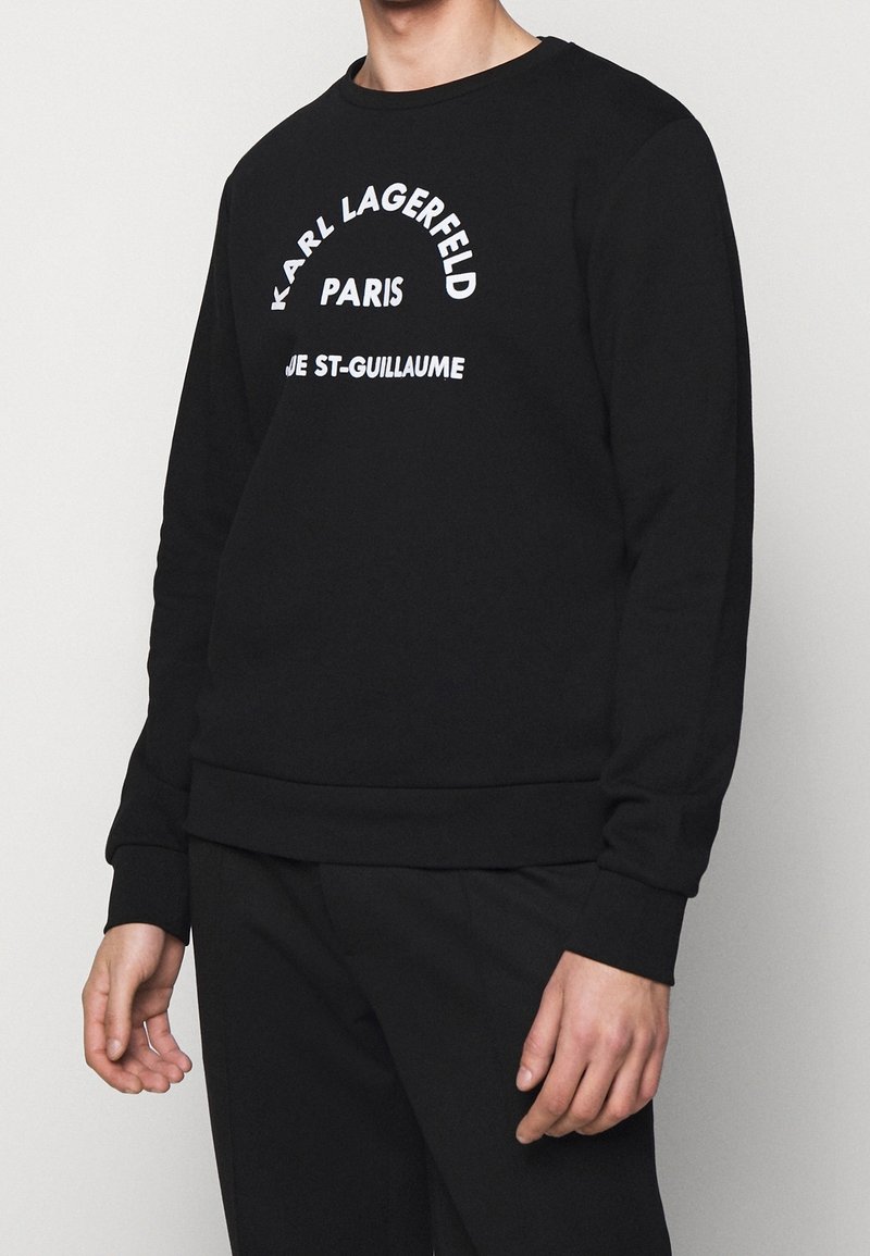 Persona con felpa nera a maniche lunghe con la scritta bianca "KARL LAGERFELD PARIS RUE ST-GUILLAUME" sul davanti, abbinata a pantaloni neri.