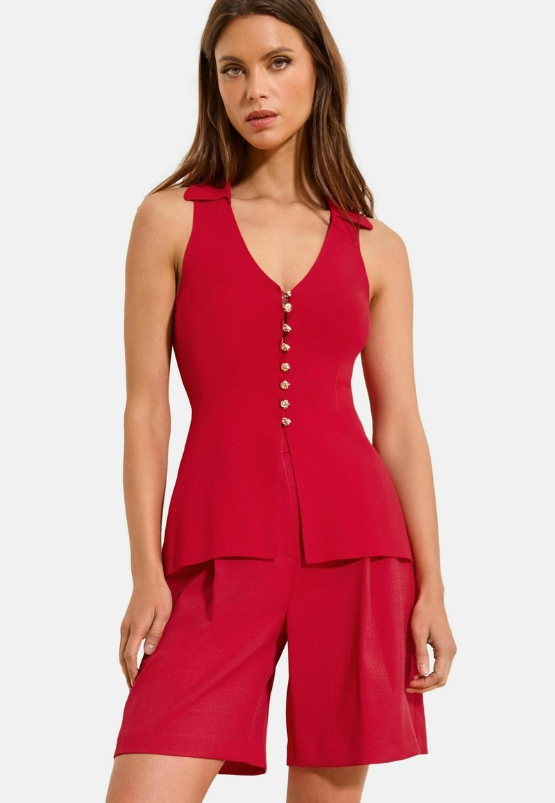 Femeie purtând un tricou roșu fără mâneci, cu guler și nasturi decorativi mici, asortat cu pantaloni scurți până la genunchi, pe fundal alb.