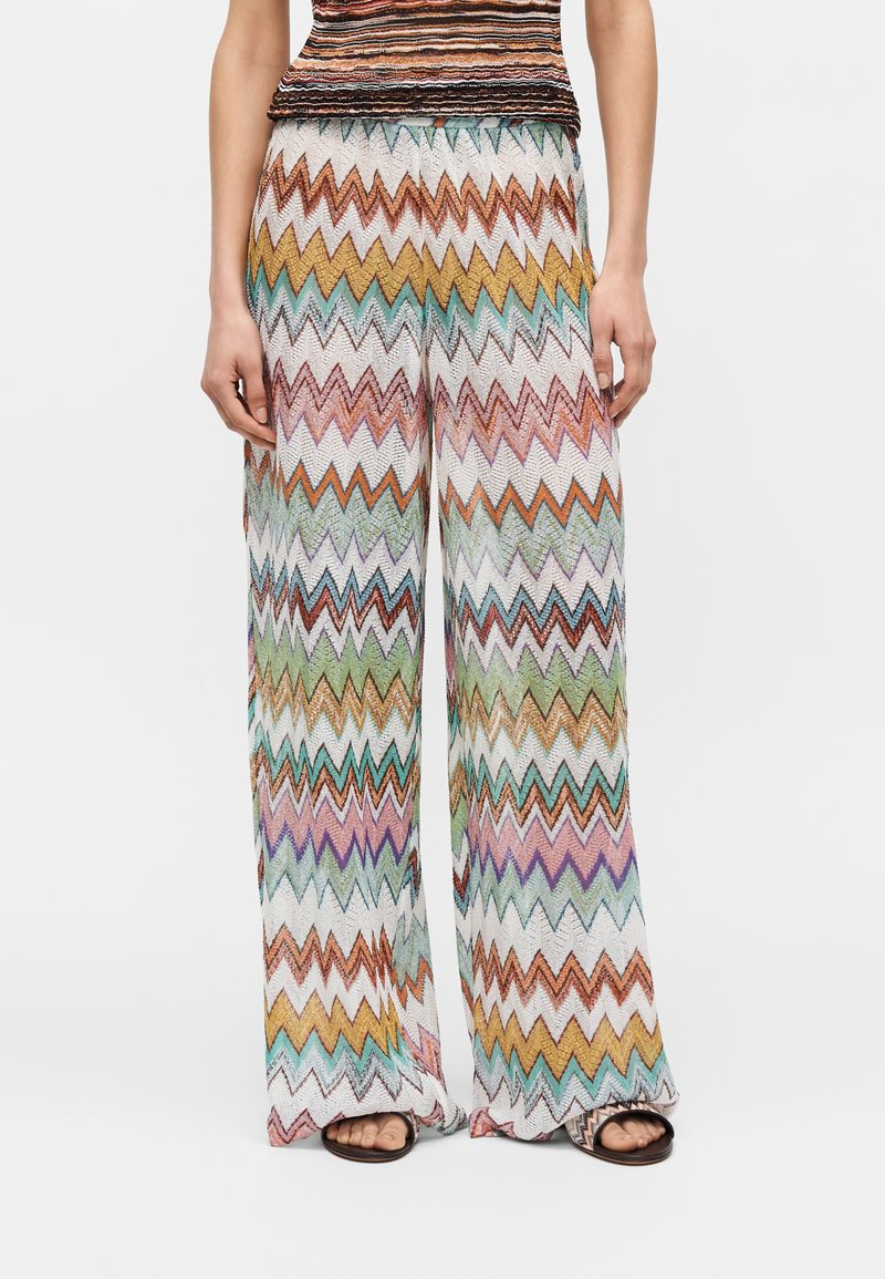 Παντελόνια με φαρδιά μπατζάκια με πολύχρωμο ζιγκ-ζαγκ σχέδιο σε αποχρώσεις πορτοκαλί, πράσινου, μωβ και λευκού, συνδυασμένα με μαύρα σανδάλια ανοιχτής μύτης.