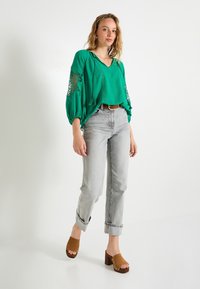 Blouse verte ample avec des manches ballon et des détails en dentelle, associée à un jean gris clair et des sandales à plateforme brunes. Texture de tissu lisse.