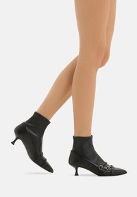 Stivali ankle neri in pelle liscia, caratterizzati da un design simile a un calzino, punte appuntite, tacco kitten e cinghie decorative con accenti metallici.