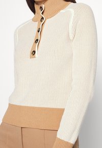 Kobieta nosząca sweter z kremowego i beżowego prążkowanego materiału z zapięciem na guziki i wysokim kołnierzem oraz pasujące beżowe spodnie.