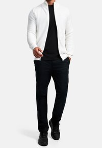 Valkoinen teksturoitu vetoketjullinen takki mustan paidan päällä, yhdistettynä mustiin housuihin ja mustiin lenkkareihin, esitettynä seisovassa hahmossa.