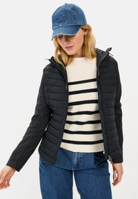 Sort pufferjakke med hætte og tekstureret krop; parret med en cremet og marineblå stribet sweater og blå denimbukser, toppet med en blå kasket.