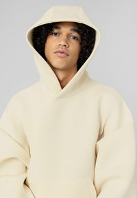 Sweat à capuche beige surdimensionné avec une grande capuche, manches longues et une poche à l'avant. Texture lisse avec peu de détails.