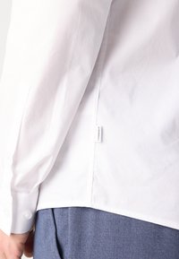 Witte lange mouw blouse met knoopmanchet, voorzien van een rechte zoom en een merklabel. Gedragen over lichtblauwe broek.